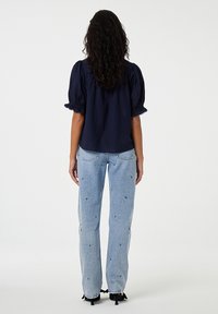 Marineblaues Oberteil mit Puffärmeln und gerafften Manschetten, kombiniert mit hellblauen Jeans mit gestickten grünen Akzenten und geradem Schnitt.