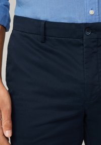 Pantalons en coton léger bleu marine avec une texture lisse, dotés d'une fermeture boutonnée et de passants de ceinture, main partiellement visible.