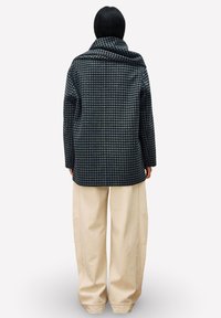 Veste à carreaux bleu marine et beige avec une coupe décontractée, présentant un large col et un pli au dos, accompagnée d'un pantalon large de couleur crème.