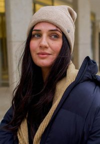 Beżowa czapka robiona na drutach z lekkim opadaniem; teksturowane wykończenie. Czarna kurtka puchowa i jasnobeżowy szalik widoczne, tworzące warstwowy look.