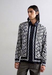 Μαύρο πουκάμισο με λευκή εκτύπωση και σχέδιο αλυσίδας; λαμπερός ύφασμα, μακριά μανίκια, κουμπωτό μπροστινό μέρος, κομψή εφαρμογή. Κύριες γραφικές λεπτομέρειες σε όλη την επιφάνεια.