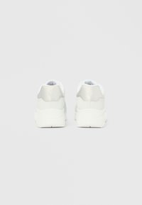 Pantofi sport albi cu partea superioară netedă din material sintetic cu finisaj mat, formă rotunjită și tălpi căptușite proeminente. Designul din spate include un accent reflectorizant.