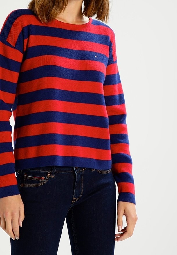 Femme portant un pull rayé horizontal rouge et bleu marine et un jean bleu foncé, debout devant un fond clair uni.