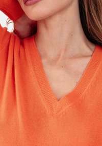 Pull à col en V de couleur orange vif, fabriqué en tissu maille doux. Présente une coupe décontractée avec des détails de coutures visibles.