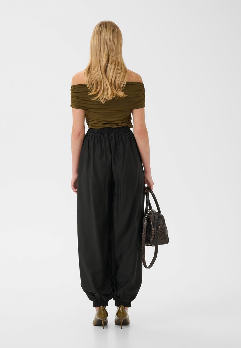 Femme aux longs cheveux blonds portant un haut olive épaules dénudées et un pantalon noir ample taille haute, tenant un sac à main noir, debout de dos.