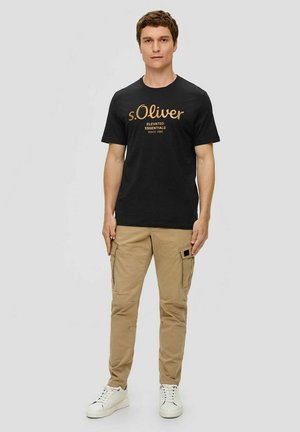 Black cotton t-shirt featuring gold text "s.Oliver Elevated Essentials Since 1969," paired with beige cargo pants and white sneakers.