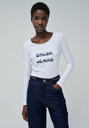 Camisa blanca de manga larga con "SALSA JEANS" en lentejuelas azul marino, combinada con pantalones de mezclilla oscuros de tiro alto con cinturón decorado con cadenas.