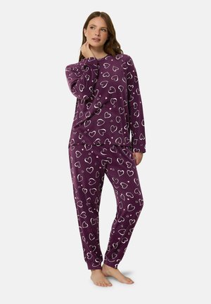 Femme debout pieds nus portant un pyjama violet à manches longues et pantalon avec des motifs de cœurs blancs sur un fond uni.