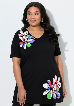 Kobieta ubrana w czarną tunikę z dekoltem w serek, ozdobioną dużymi, kolorowymi kwiatowymi wzorami na ramieniu i biodrze, stojąca na tle jednolitego, białego tła.