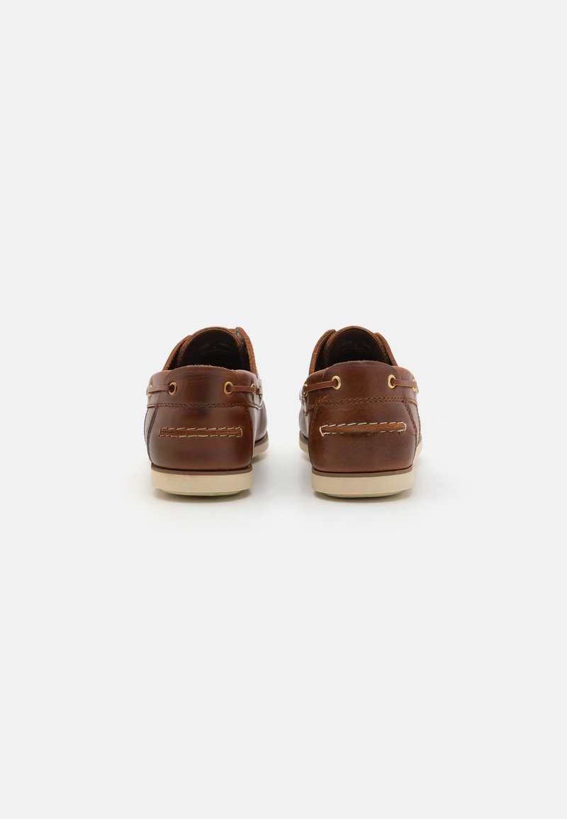 Chaussures de bateau en cuir marron avec des coutures beiges, embout rond et semelles en caoutchouc. Présente deux oeillets pour les lacets de chaque côté.