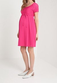 Rosa Umstandskleid mit kurzen Spitzenärmeln, Taillenband und plissiertem Rock. Kombiniert mit weißen Slip-On-Schuhen. Glatte Stoffstruktur.