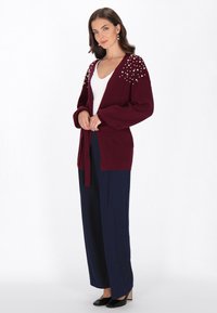 Bordowy kardigan z perłowymi akcentami na ramionach, otwarty z przodu, z prążkowanej tekstury, w połączeniu z granatowymi spodniami o szerokich nogawkach i czarnymi butami.