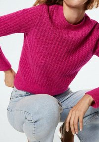 Vrouw in een geklede felroze trui en lichtblauwe jeans, hurkend met één hand op haar heup en de andere op haar knie.