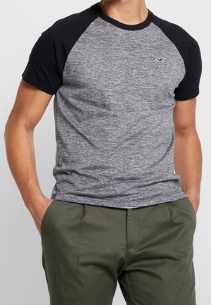 Graues Melange-T-Shirt mit kurzen Ärmeln, schwarzen Raglanärmeln und kleinem Logo auf der Brust, kombiniert mit olivgrünen Hosen.