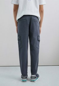Pantalones cargo grises con bolsillos laterales, corte recto y detalles de costura visibles. Llevados con zapatillas oscuras. Vista trasera contra un fondo sencillo.