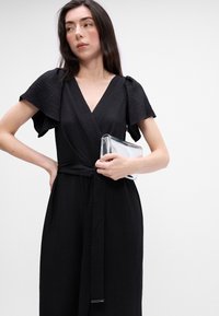 Rochie neagră cu mâneci fluture, din țesătură texturată și talie beltată. Ținând o geantă de mână argintie lucioasă.