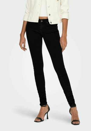 Femme portant un jean skinny noir, des sandales à talons noirs, un top court blanc et une veste blanche, debout devant un fond clair.
