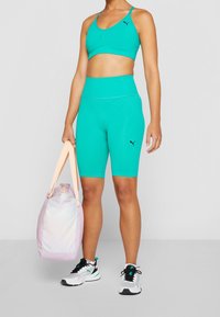 Femme portant un bras de sport turquoise et un short avec des logos Puma, tenant un sac de sport rose clair et portant des baskets blanches et noires.