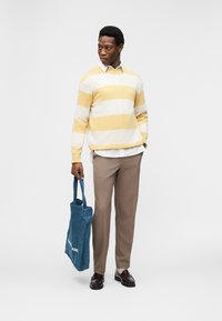Homme portant un pull à rayures jaunes et blanches, un pantalon beige et des chaussures marron, tenant un sac fourre-tout bleu, regardant vers sa gauche.