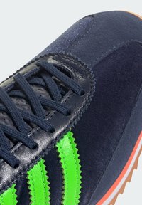 Adidași din suede bleumarin închis cu dungi sintetice verzi, șireturi negre, finisaj texturat și talpă din cauciuc de tip gumă. Au un design retro.