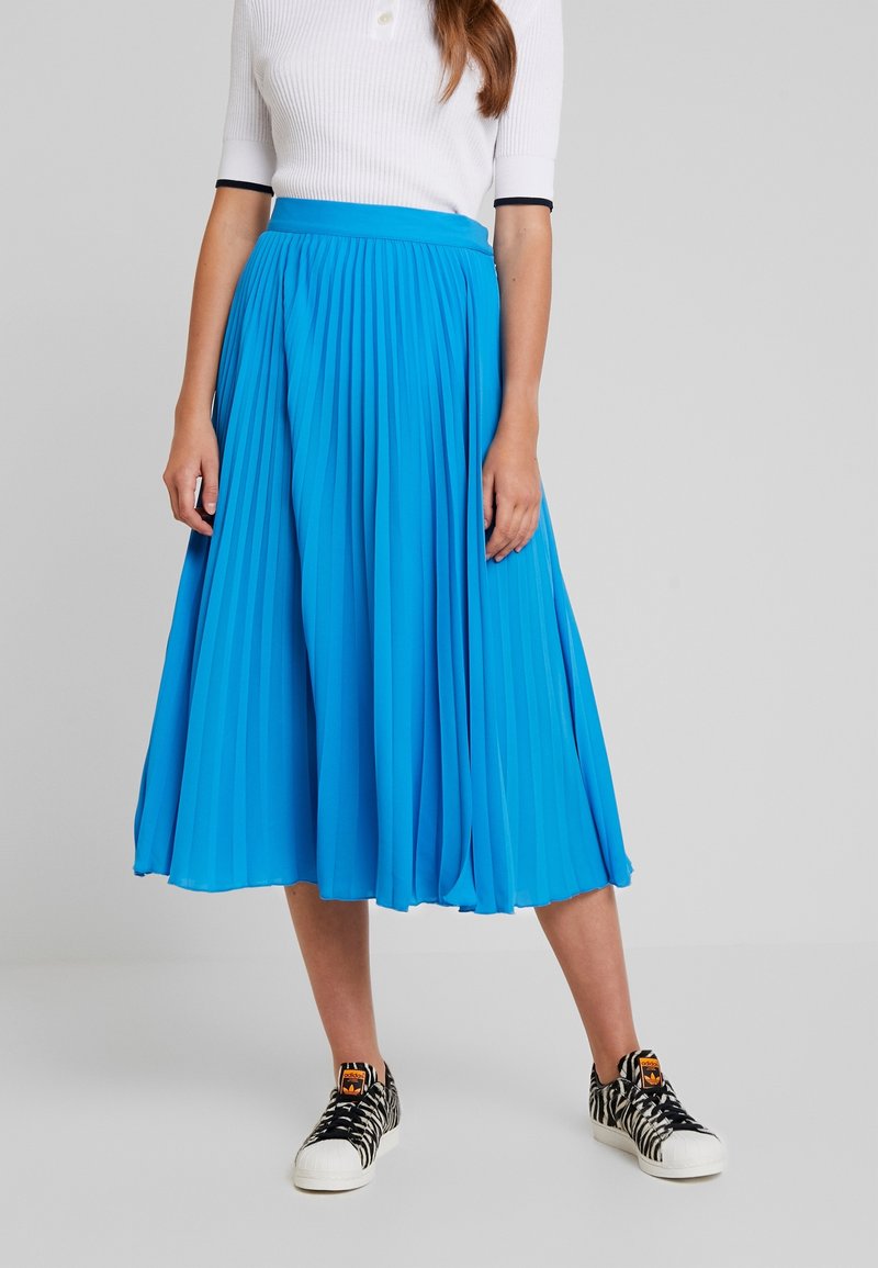 Femme portant un haut blanc à manches courtes, une jupe midi plissée bleu vif et des baskets à motifs noir et blanc, debout sur un fond gris.