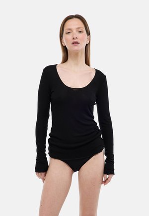 Femme debout portant un haut noir à manches longues avec un col en U et une culotte noire assortie, sur un fond clair uni.