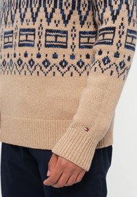 Pull en laine beige tricoté avec des motifs géométriques marins, des poignets côtelés et un accent logo sur la manche. Texture lisse et coupe décontractée.