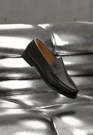 Schwarzer Loafer aus Leder mit Krokodilprägung, der über einem glänzenden silberfarbenen, gesteppte Sofa schwebt und Details sowie Textur des Schuhs im Nahaufnahme zeigt.