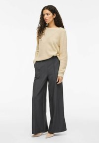 Pull en tricot beige avec un col large associé à un pantalon large taille haute en charbon avec des plis devant.