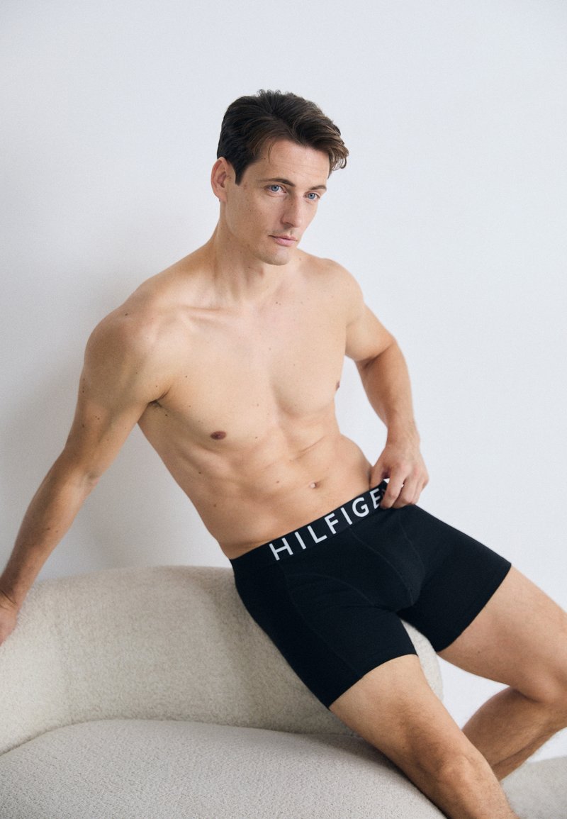 Skjorteløs mann med mørkt hår iført svarte Tommy Hilfiger boxershorts, sittende på en teksturert kremfarget sofa mot en enkel hvit vegg.