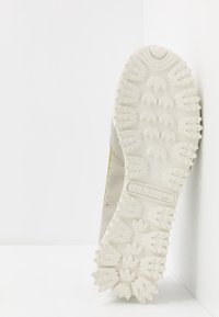 Ljusgrå träningsskor med en texturerad, vit gummisula som har ett mönstrat grepp. Accentuerad med en liten gul rand.