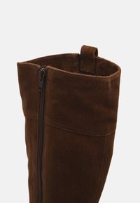 Bota de ante marrón con cierre de cremallera en el lateral, con una textura suave y un borde superior redondeado. Incluye una lengüeta para facilitar su colocación.