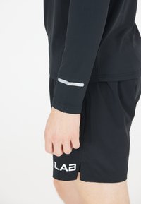 Sort langærmet sportsbluse med reflekterende detaljer på ærmet, parret med sorte shorts med et hvidt logo ved kanten.