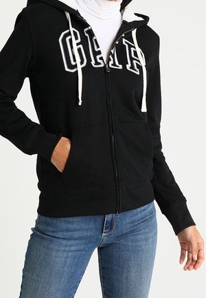 Person wearing a black zip-up hoodie with white "GAP" letters and white drawstrings, paired with blue jeans, hand in pocket.