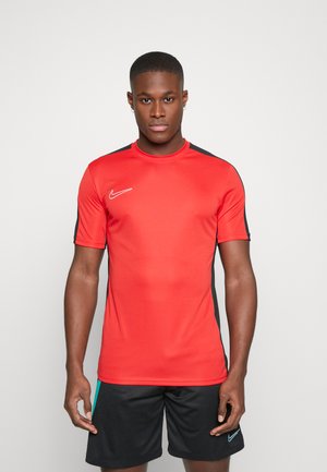 Camiseta deportiva roja de manga corta, con un cuello redondo, detalles negros en los laterales y un logo de Nike blanco en el pecho. Llevada con pantalones cortos negros.
