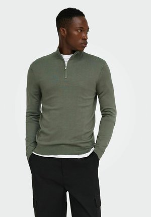 Ung mand iført en olivengrøn sweater med kvart lynlås over en hvid skjorte og sorte bukser, ser mod højre med hænderne i lommerne.