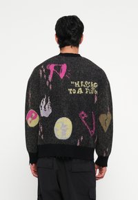 Jersey negro de punto con diseños gráficos coloridos que incluyen corazones, llamas y varios símbolos. Dobladillo y puños deshilachados. Corte relajado.