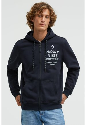 Young man with curly hair wearing a navy blue zip-up hoodie with “Beach Vibes” text and light blue jeans, standing against a plain background.