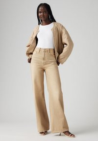 Giacca bomber trapuntata beige sopra una maglietta bianca, abbinata a jeans a vita alta beige chiaro a gamba larga e tacchi aperti color cammello.
