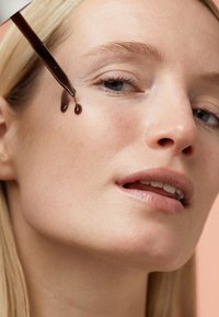 Tropfflasche gibt eine dunkle Flüssigkeit auf die Haut nahe dem Auge ab. Glatte Haut mit sanfter Beleuchtung, vor einem pastellfarbenen Hintergrund.