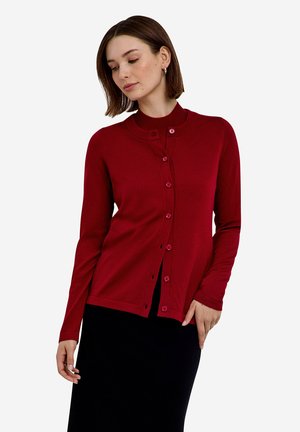 Frau mit kurzen braunen Haaren, die eine rote, geknöpfte Strickjacke über einem schwarzen Rock trägt, schaut nach unten, mit einer Hand nahe der Hüfte.