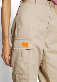 Beige cargo kalhoty vyrobené z odolné tkaniny, s bočními kapsami a oranžovou nášivkou "OBEY" na přední kapse, s volným střihem.