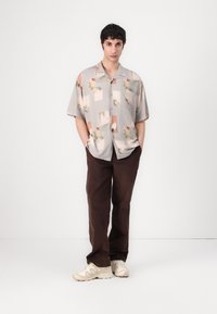 Cămașă cu mânecă scurtă, cu nasturi, având o bază gri deschis, prezentând un model floral și geometric. Asortată cu pantaloni maronii și adidași bej.