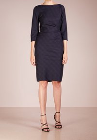 Femme portant une robe noire à mi-longueur avec des manches trois-quarts et des sandales à talons hauts noires à lanières, se tenant sur un sol beige.