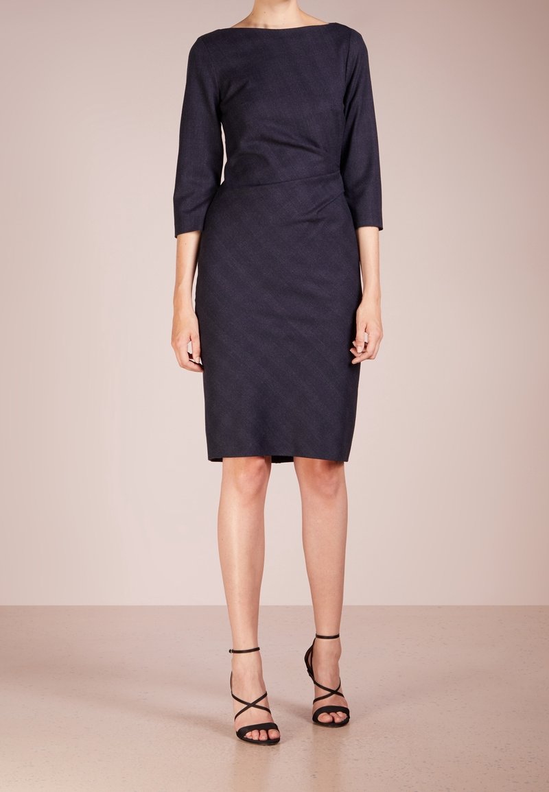 Femme portant une robe noire à mi-longueur avec des manches trois-quarts et des sandales à talons hauts noires à lanières, se tenant sur un sol beige.