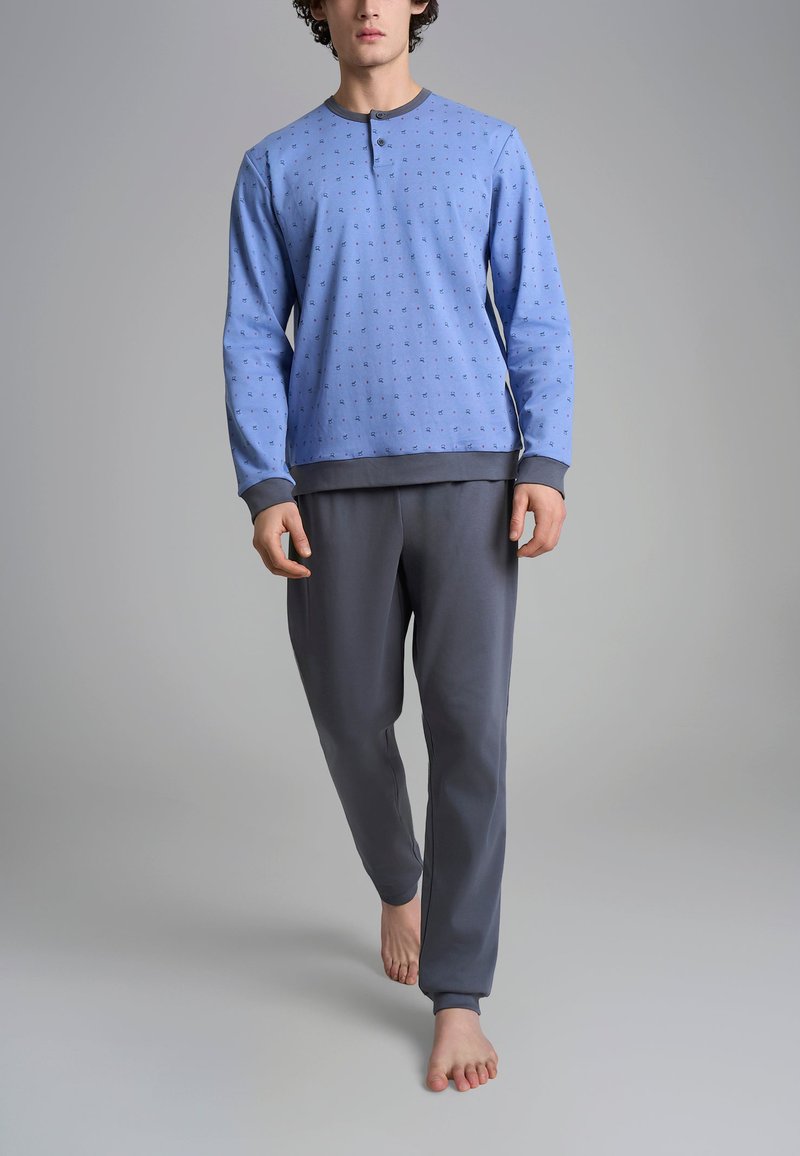 Maglietta blu a maniche lunghe con piccoli motivi e scollo rotondo, abbinata a pantaloni jogger grigi, entrambi in tessuto morbido. Modella in piedi a piedi nudi.