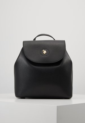 Mochila de cuero negro con asa superior y cierre de solapa que presenta un pequeño emblema dorado sobre un pedestal blanco.