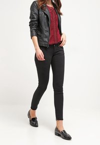 Svart läderjacka över en bordeauxfärgad blus med spetsdetaljer, kombinerad med svarta åtsittande jeans och glänsande svarta loafers, vilket skapar en stilren och koordinerad outfit.