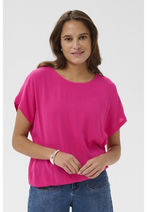 Femme aux cheveux châtain moyen portant un haut rose vif à manches courtes et un jean bleu, debout les mains légèrement jointes.