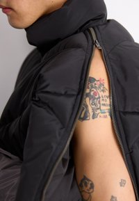 Črn puhovka z visokim ovratnikom, z odpiranjem na strani. Ima gladko, mat zunanjost, pod katero se vidi tatuiran roka.